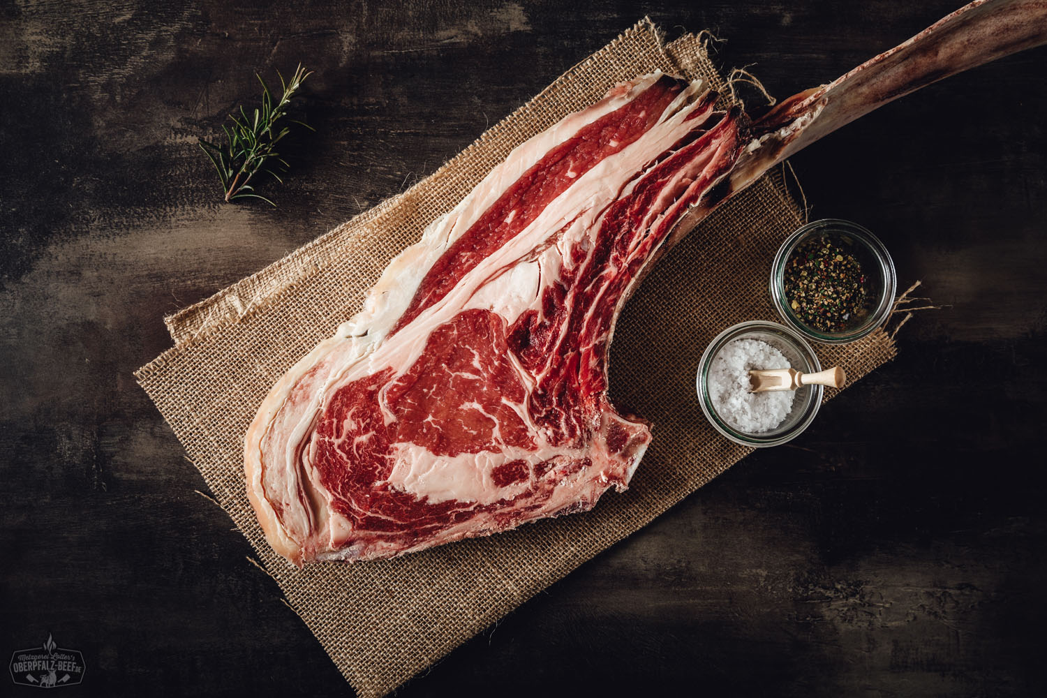 Dry Aged Tomahawk Steak von der alten Kuh – Premium Beef aus der Oberpfalz Tomahawk-Steak Dry Aged von der alten Kuh, roh und mit Knochen, auf dunklem Holzbrett angerichtet, mit grobem Meersalz, Rosmarinzweigen und Knoblauch garniert – hochwertiges Rindfleisch mit intensiver Marmorierung für Grillen und Feinschmecker.
