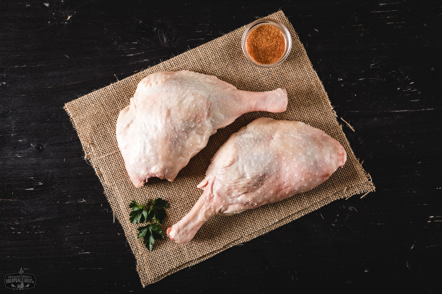 Keulen von der Oberpfälzer Bauernente frisch und regional Rohes Entenfleisch von der Oberpfälzer Bauernente, auf einem dunklen Schneidebrett angerichtet, flankiert von frischen Kräutern, Zwiebeln, Knoblauch, Salzflocken und grobem Pfeffer – hochwertige Zutaten für traditionelles Geflügelgericht aus Bayern.