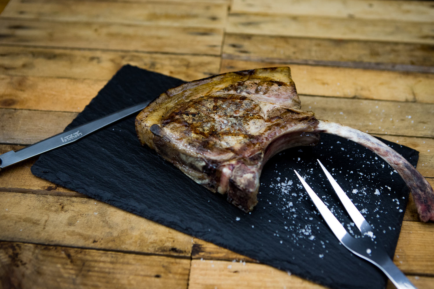 Tomahawk-Steak vom Oberpfälzer Landschwein – Dry Aged Fleischgenuss Tomahawk-Steak vom Oberpfälzer Landschwein, dry-aged: Rohes, marmoriertes Schweinesteak mit langem Knochen, auf schwarzem Steinbrett angerichtet und mit Meersalz, Kräutern und Gewürzen garniert, rustikale Darstellung für BBQ oder Premium-Fleischgenuss.