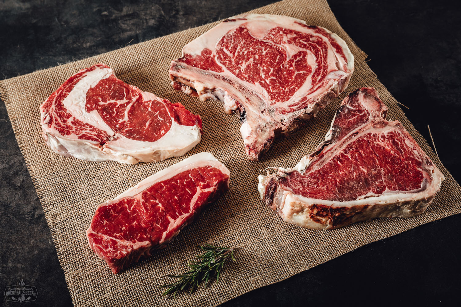 Drei vakuumverpackte, unterschiedlich große Dry-Aged Steaks liegen nebeneinander auf einem dunklen Untergrund; gut sichtbare Marmorierung des Fleisches, daneben ein Oberpfalz Beef Etikett mit Logo – hochwertiges Rindfleischpaket aus regionaler Aufzucht.