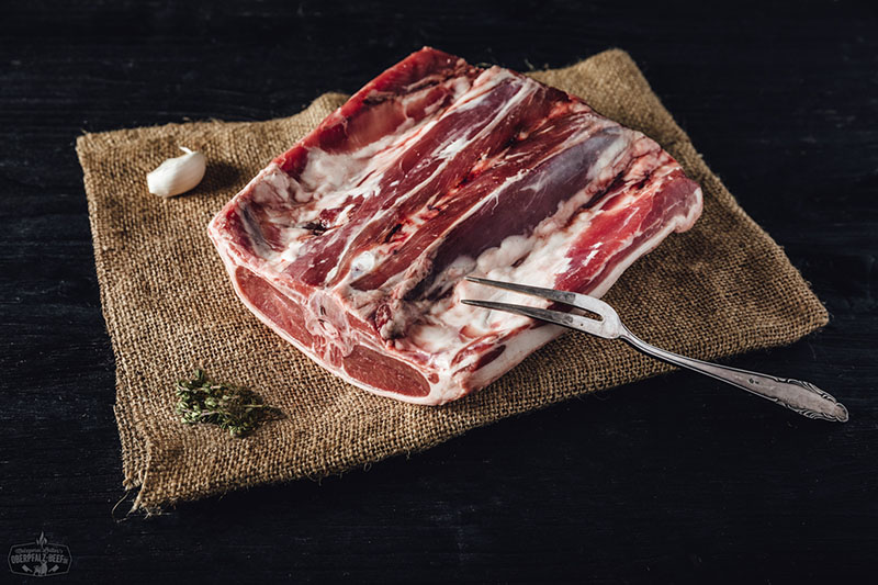 Ganzes Lammrücken-Kotelett vom Juradistl-Lamm, roh und mit Knochen, auf Holzbrett angerichtet; saftiges, marmoriertes Fleisch mit rosiger Farbe, natürliche Fettschicht sichtbar, rustikale Darstellung für Fleischliebhaber und Gourmetküche.