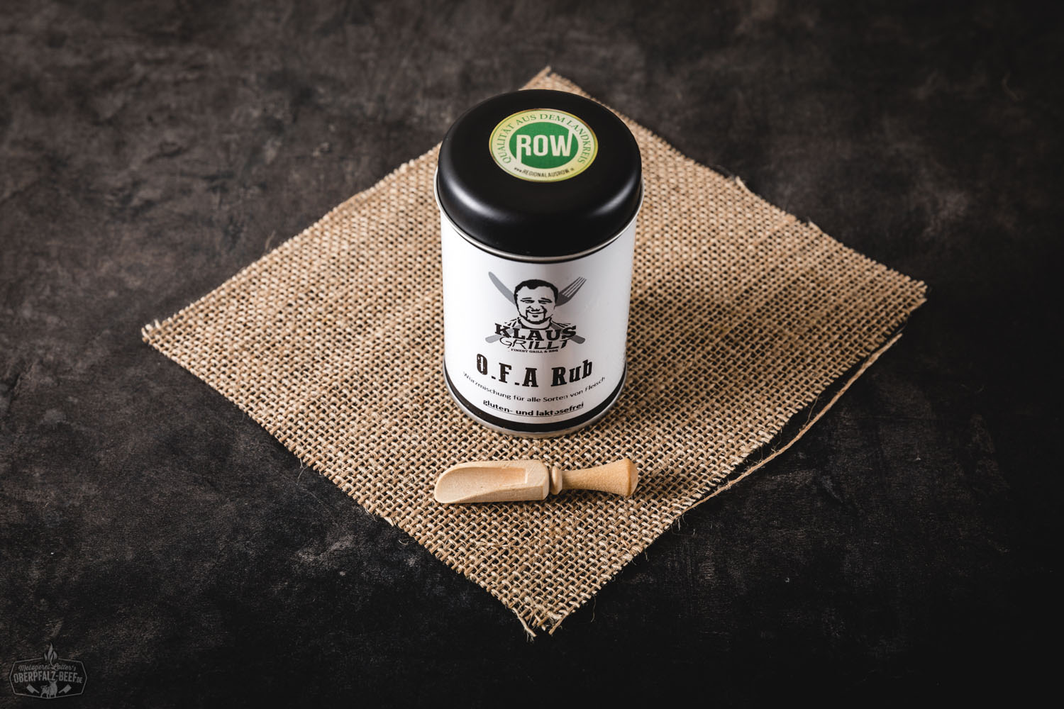 Mann grillt Fleisch auf Holzkohlegrill, im Vordergrund steht eine geöffnete Gewürzdose mit der Aufschrift „OFA Rub“, rauchiger Grillduft steigt auf – sommerliches BBQ-Szenerie mit Fokus auf Grillgewürz von Oberpfalz Beef.