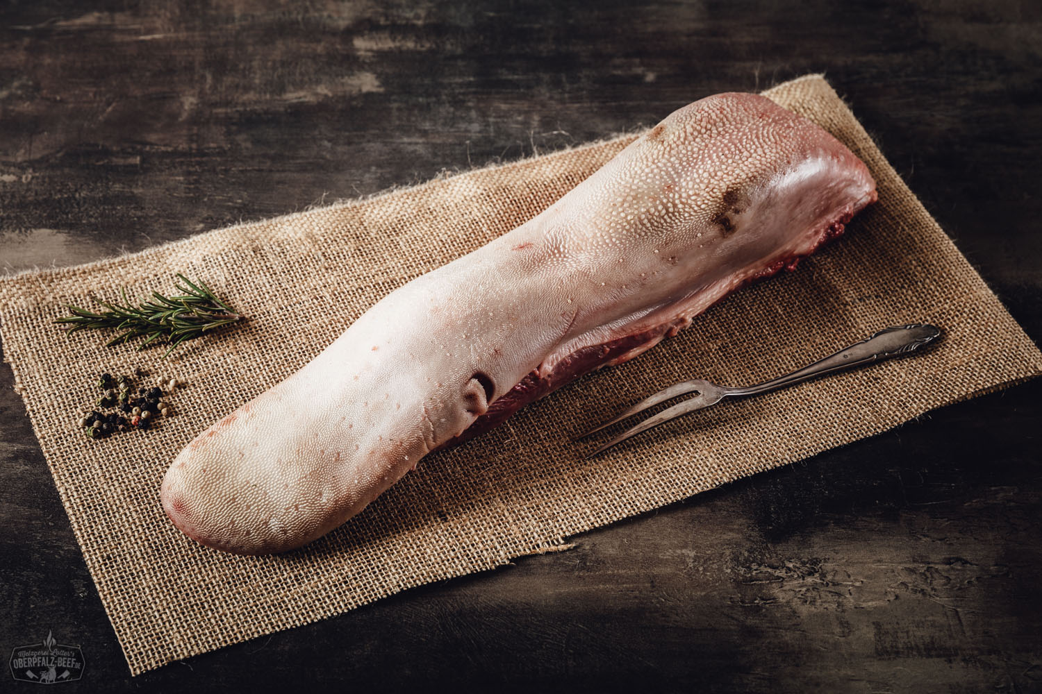 Rohes Stück Rinderzunge auf einem dunklen Untergrund, umgeben von frischen Zutaten wie Zwiebeln, Petersilie, Knoblauchzehen und Gewürzen; ideal für rustikale Fleischgerichte und traditionelle bayerische Küche.