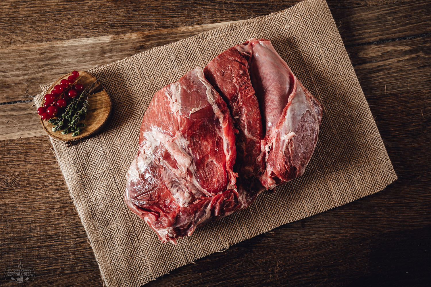 Roher Wildschwein-Schulterbraten ohne Knochen aus der Oberpfalz auf dunklem Untergrund, fein marmoriert, angerichtet mit Rosmarin, Knoblauchzehen und bunten Pfefferkörnern – hochwertiges Wildfleisch für nachhaltigen Fleischgenuss.