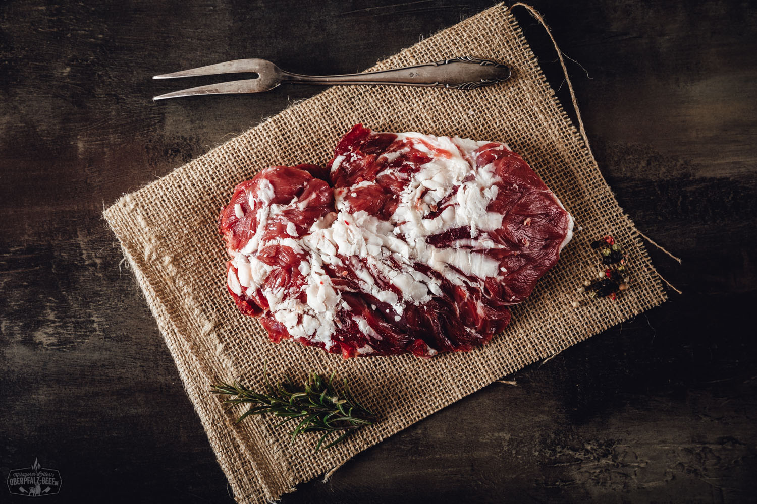 Spider-Steak Wet Aged vom Oberpfalz-Rind – Zartes Rindfleisch in Premium-Qualität Rohes Spider Steak vom Oberpfalz-Rind, wet-aged gereift, liegt auf dunklem Untergrund mit sichtbarer feiner Marmorierung; umgeben von grobem Meersalz, schwarzem Pfeffer und frischen Kräutern – hochwertiger Zuschnitt für Steakliebhaber.