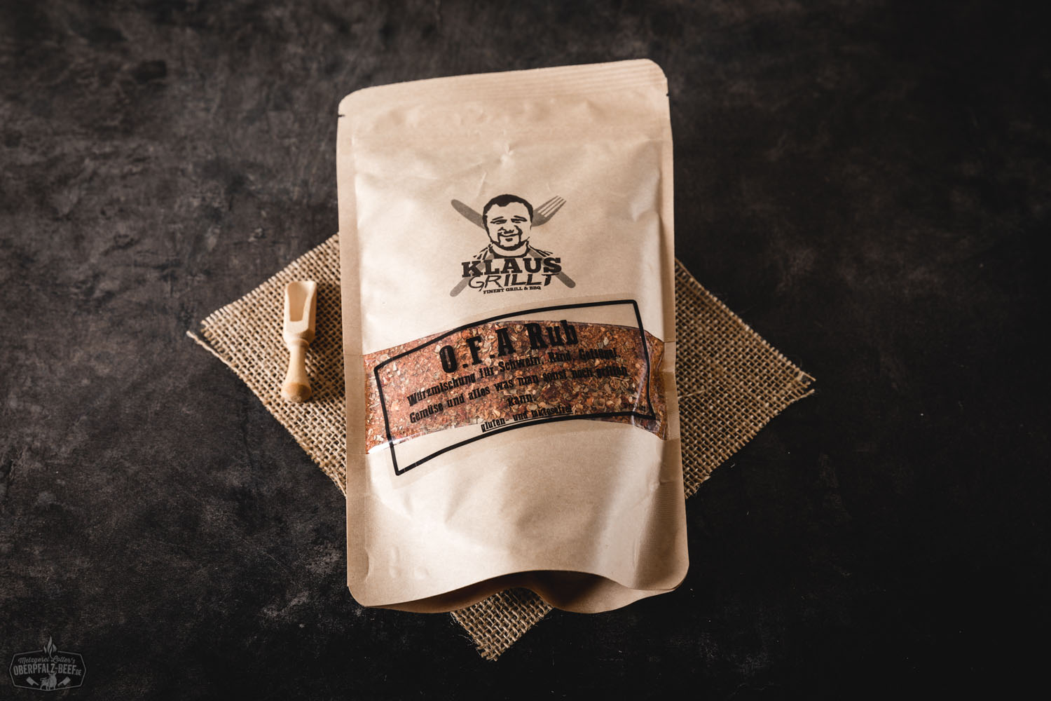OFA Rub Gewürzmischung im Beutel – BBQ Grillgewürz von Klaus grillt Mann steht im Garten neben einem Grill und hält lächelnd einen Beutel mit OFA BBQ Rub von Oberpfalz Beef in die Kamera; der Gewürzbeutel ist deutlich lesbar, Sonne scheint, entspannte Grillatmosphäre im Freien.