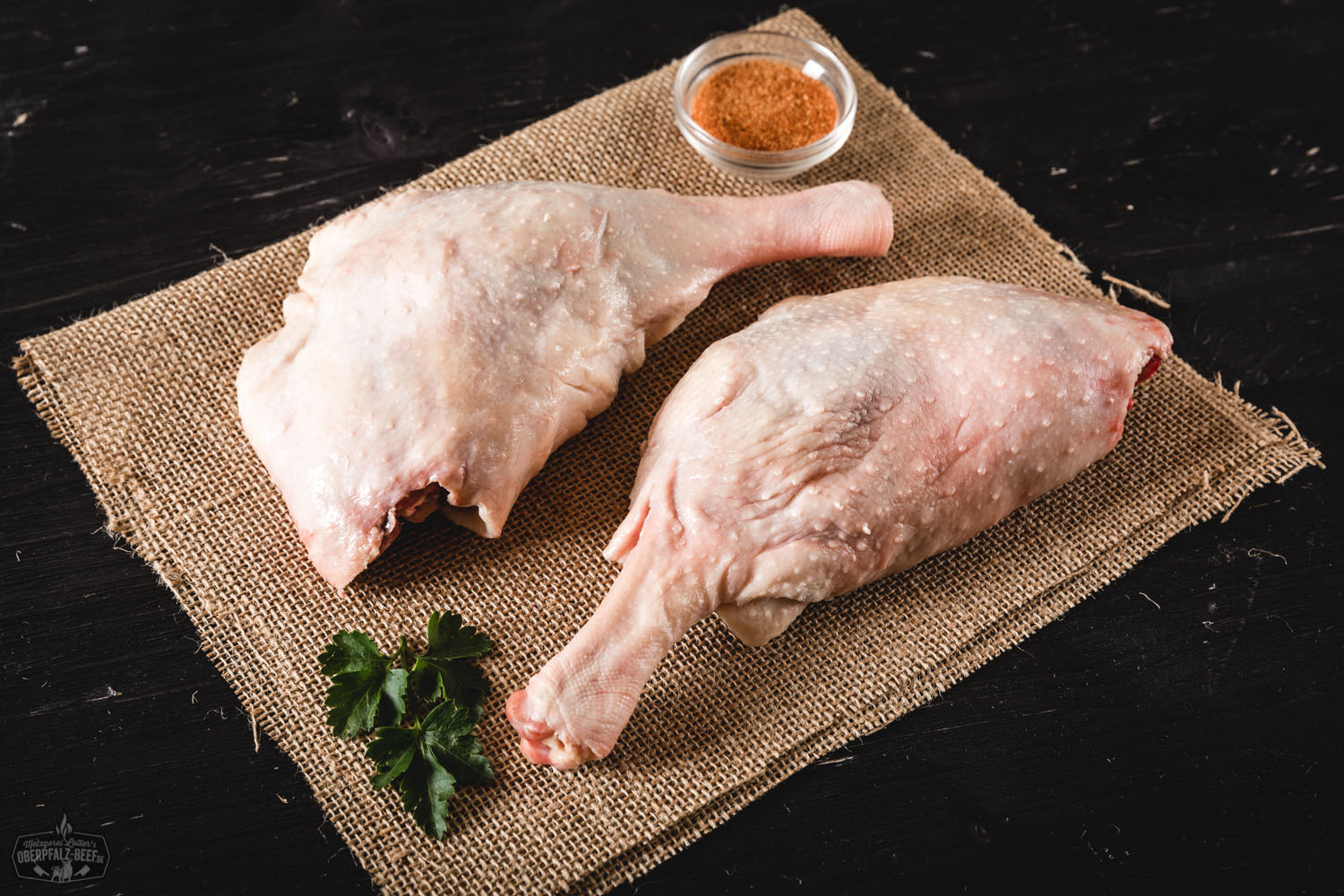 Zwei goldbraun gebratene Gänsekeulen der Oberpfälzer Bauerngans liegen auf einem rustikalen Holzbrett, garniert mit frischen Kräutern und umgeben von Bratensaft; im Hintergrund sind ein Messer, eine Holzgabel und herbstliche Dekoelemente zu sehen.