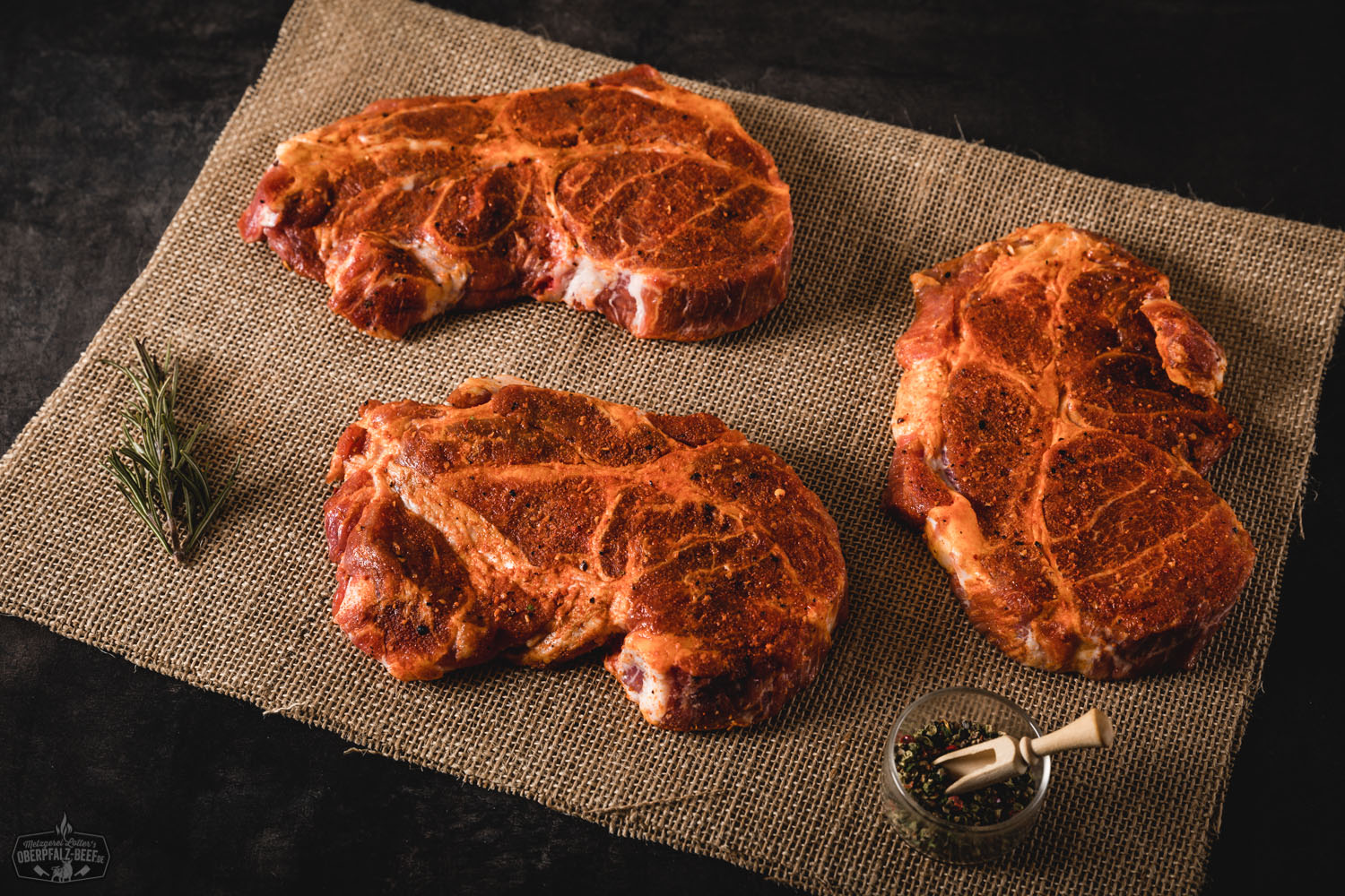 Zwei saftig gewürzte Rinderhalssteaks liegen nebeneinander auf einem dunklen Schneidebrett, umgeben von grobem Salz, buntem Pfeffer, frischen Kräutern und Knoblauchzehen – ideal für Grillfans und Fleischliebhaber.