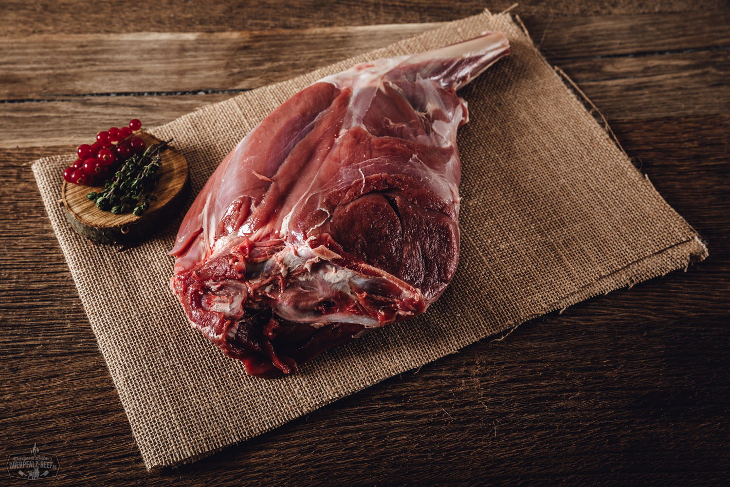 Roher Rehschulterbraten mit Knochen vom Oberpfälzer Reh auf dunklem Holzbrett, umgeben von frischen Rosmarinzweigen, Wacholderbeeren und grobem Salz – hochwertiges Wildfleisch für Braten oder Schmorgerichte.