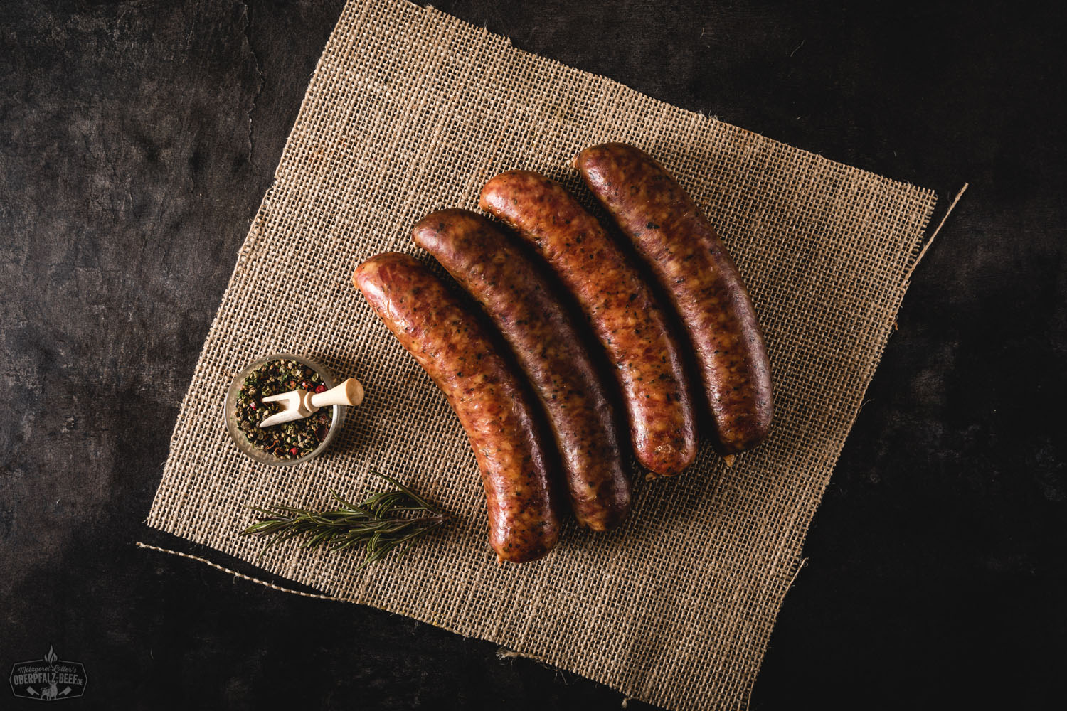 Bauernseufzer Wurst frisch gegrillt auf rustikalem Holzbrett mit Senf und Brot Vier grob gekörnte, gebräunte Bauernseufzer-Würste aus Schweinefleisch liegen nebeneinander auf einem Holzbrett, umgeben von frischen Kräutern, Zwiebelringen und buntem Pfeffer – rustikal angerichtet für eine herzhafte Brotzeit.