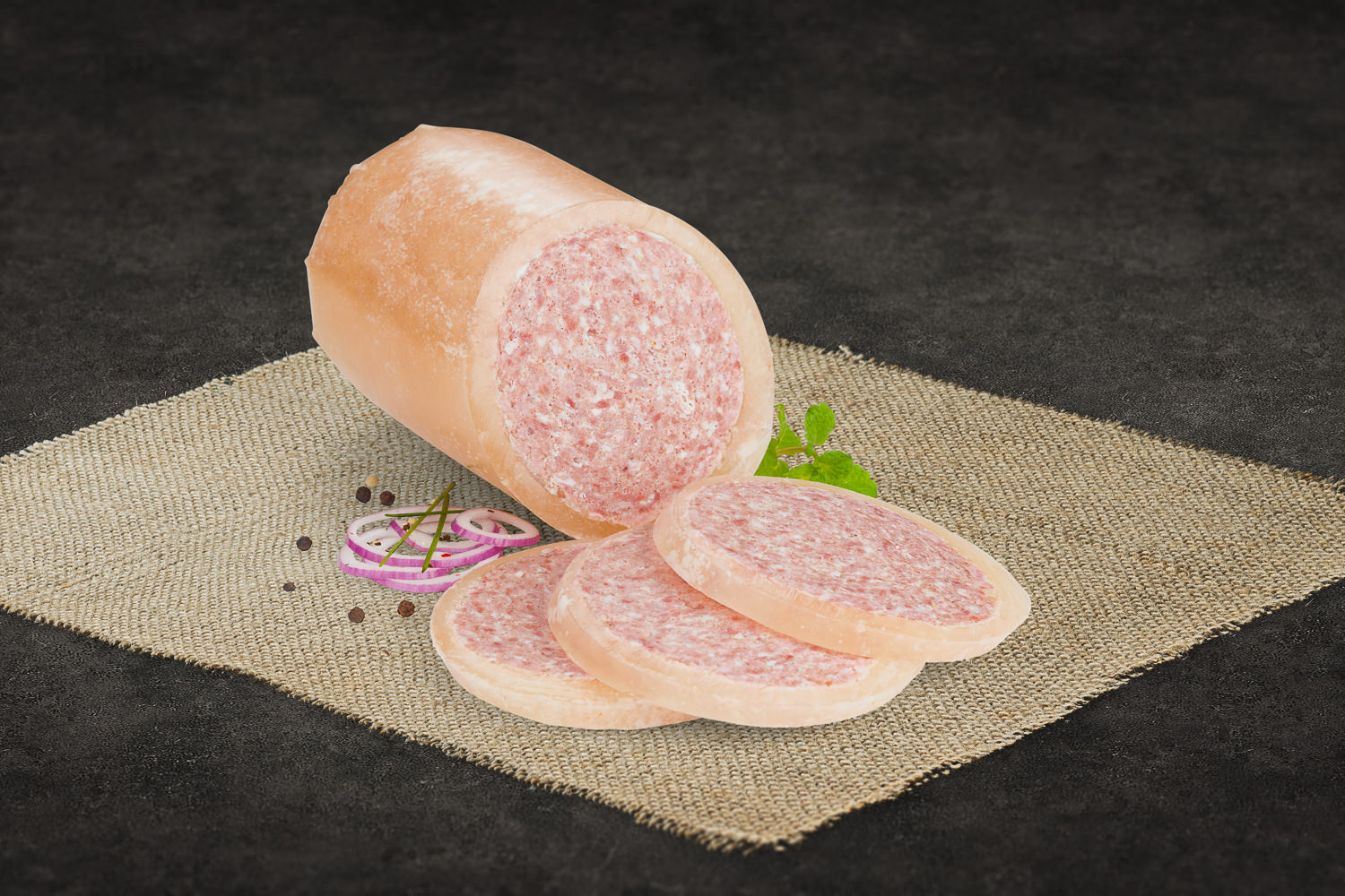 Mehrere Scheiben einer aufgeschnittenen Peppadew-Wurst mit sichtbaren roten Paprikastückchen, angerichtet auf einem dunklen Schneidebrett aus Holz; im Hintergrund ganze Peppadew-Schoten – hochwertige Gourmet-Wurst mit pikanter Note.