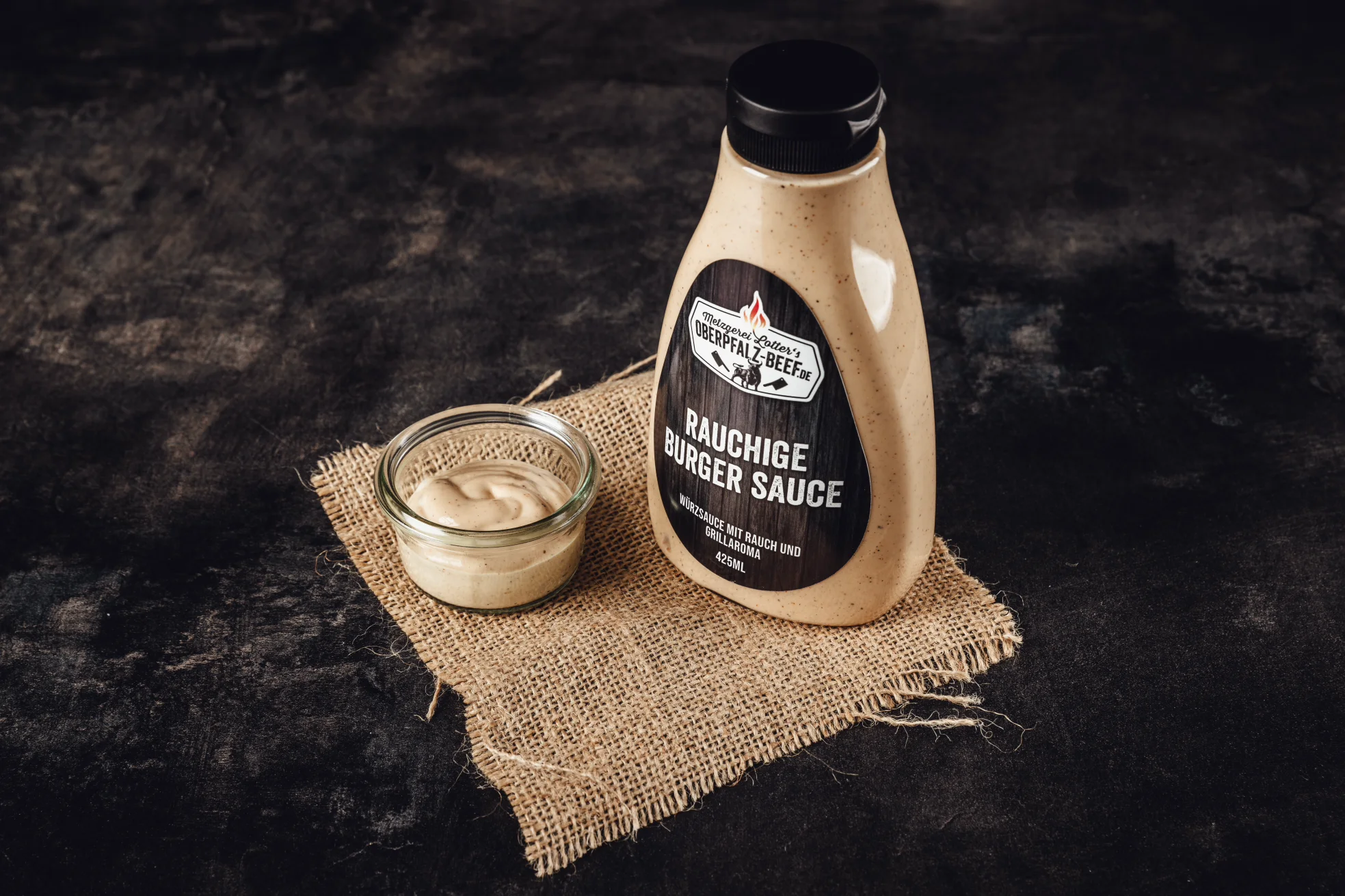 Rauchige Burger Soße in Glasflasche mit Etikett – Oberpfalz Beef BBQ-Sauce Grillteller mit saftig gegrilltem Rindfleisch, Bratwurst und Maiskolben auf rustikalem Holzbrett, serviert mit Grillzange und Kräutern – Oberpfalz Beef Werbung für Fleisch-Gutschein im BBQ-Stil.